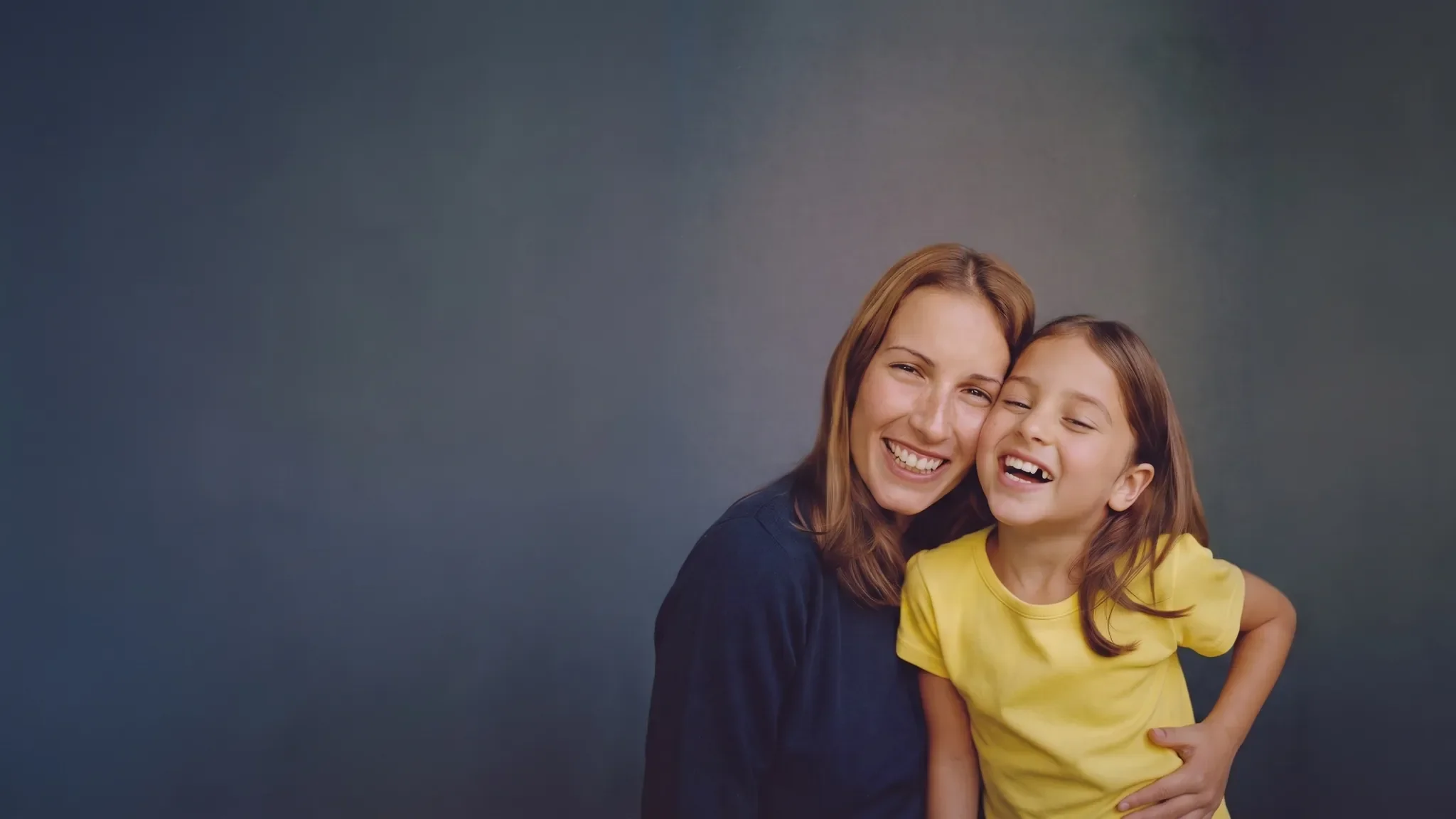 Mother and young daughter laughing cheek to cheek against a plain studio backdrop